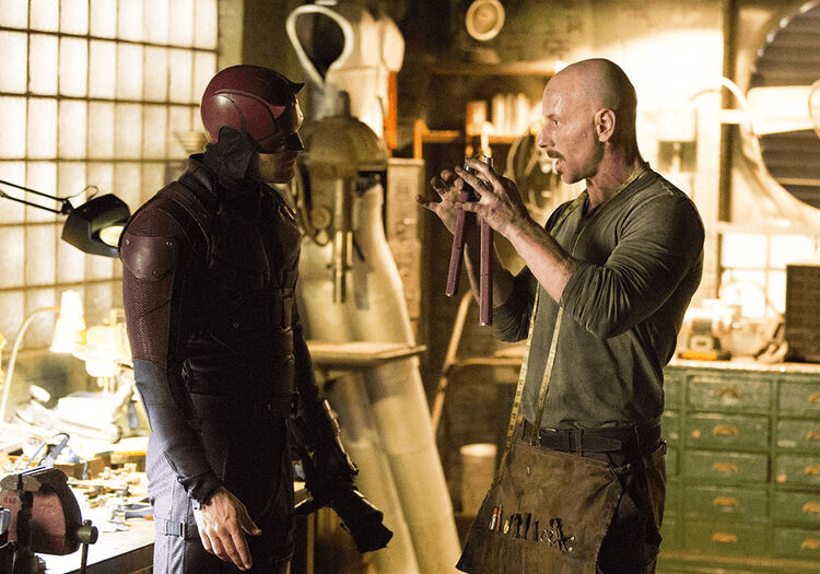 Daredevil in his red suit talking to two people inside a workshop