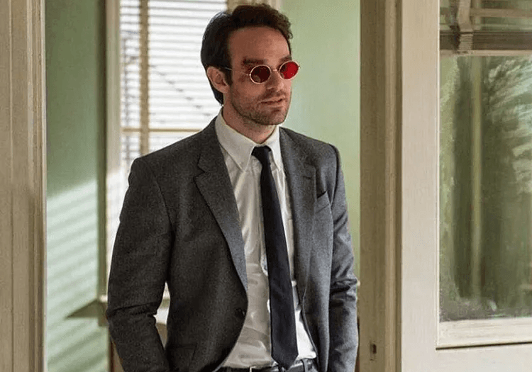 Matt Murdock in a grey suit and red tinted glasses standing by a window